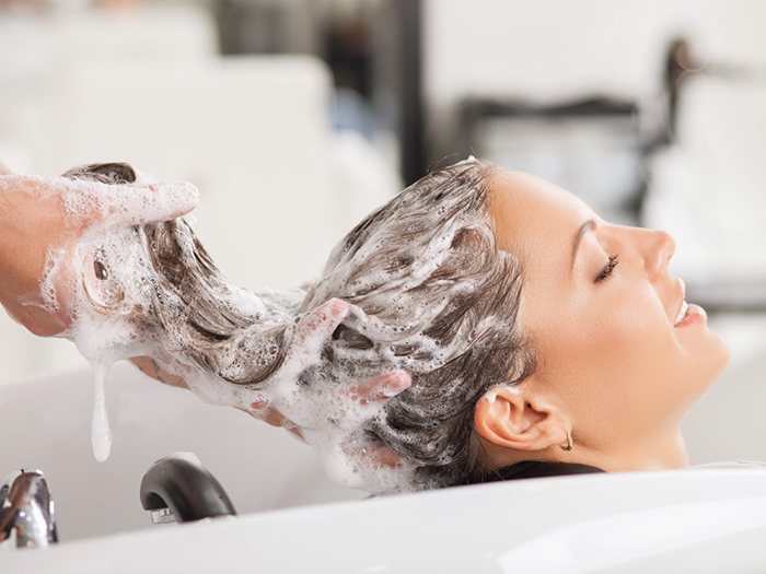 Close up of face of beautiful woman is getting a hairwash by a hairdresser. She is smiling. Her eyes are closed with relaxation. The beautician is making foam from shampoo
